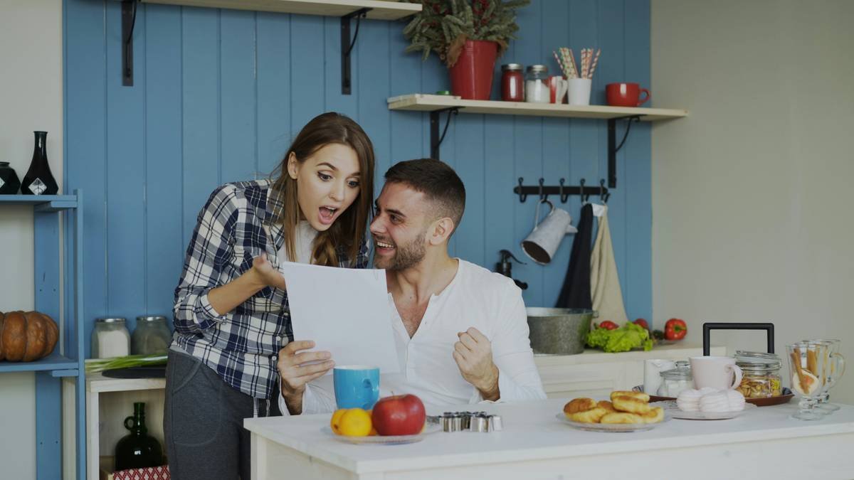 Attractive happy couple recieve good news unfolding letter in the kitchen while have breakfast at home early morning
